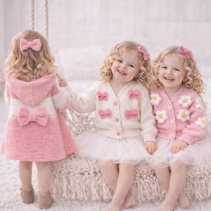 May include: Three young children in knitted outfits. One wears a pink coat with a bow, the others cardigans with bows and flower details. All have matching hair bows. The outfits are in shades of pink and white.