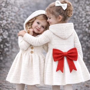 May include: Two young girls in matching cream-colored hooded dresses with a red bow on the back. The dresses have a button closure and are paired with white tights. One girl has a white bow in her hair.
