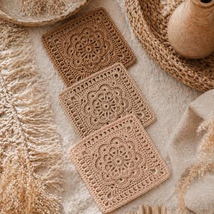 Puede incluir: Tres posavasos cuadrados de ganchillo en tonos beige y marrón. Cada posavasos presenta un diseño floral central y un borde decorativo. Los posavasos están dispuestos sobre una superficie blanca, con una cesta tejida y un jarrón de cerámica en el fondo.