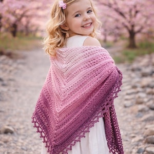 May include: A crocheted shawl in shades of pink and purple, draped over the shoulders. The shawl has a V-shape and a scalloped edge. The background features a path and trees with pink blossoms.