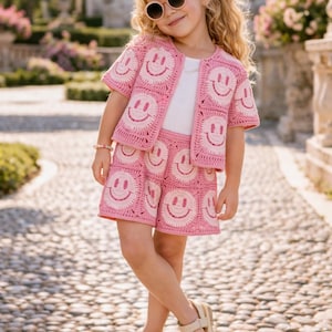 Könnte beinhalten: Ein Kind trägt ein rosa Häkel-Outfit mit Smiley-Gesichtern. Das Outfit besteht aus einer kurzärmeligen Jacke und Shorts. Das Kind trägt auch eine Sonnenbrille und Sandalen. Das Outfit ist aus hellrosa Garn gefertigt.