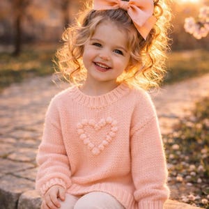 May include: A smiling child with curly blonde hair and a large peach-coloured bow. She wears a light pink jumper with a heart design and light-coloured trousers. The background is blurred with warm tones.