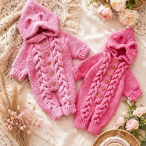 Peut inclure: Deux barboteuses roses en tricot pour bébé, avec capuches et boutons en bois. Les barboteuses présentent un motif torsadé sur le devant et de petits détails d'oreilles sur la capuche. Les vêtements sont présentés sur une surface crème avec des accents floraux.