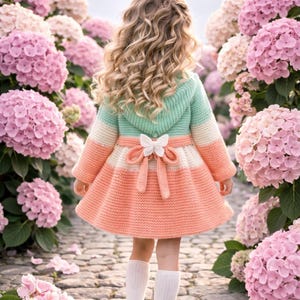 Könnte beinhalten: Ein junges Mädchen in einem pastellfarbenen Strickkleid mit einer Schmetterlingsschleife, steht in einem Garten mit rosa und weißen Hortensien. Das Kleid hat eine mintgrüne Kapuze und Ärmel mit Pfirsich- und Cremeakzenten.