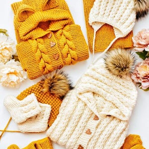 May include: A selection of hand-knitted winter clothing in mustard yellow and cream. The set includes a hooded jacket with wooden buttons, a scarf, a hat with a faux fur pom-pom, and matching mittens. The items are displayed on a white surface.