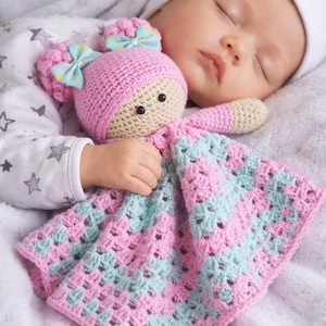 Puede incluir: Un bebé dormido abraza una muñeca y una manta de ganchillo hechas a mano. La muñeca tiene un gorro rosa con lazos y una cara beige. La manta es rosa y azul claro. El bebé lleva un gorro blanco y un body con estampado de estrellas.