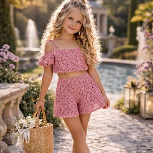 May include: A girl in a pink crochet crop top and shorts set, holding a woven basket with white flowers. The outfit is a light pink colour. The background shows a fountain and garden setting.