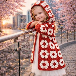 Könnte beinhalten: Ein rot-weißer gehäkelter Kapuzenmantel mit Blumenmuster. Der Mantel wird über einem weißen Kleid getragen. Der Hintergrund zeigt eine Skyline und rosa blühende Bäume. Der Mantel ist ein einzigartiges handgefertigtes Produkt.