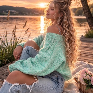 May include: A woman wearing a light green off-the-shoulder sweater and ripped blue jeans sits on a wooden dock. The background features a lake and a sunset. A picnic basket with pink flowers and sunglasses are nearby.