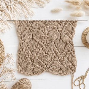 May include: A tan knitted fabric with a leaf-like pattern is displayed on a white wooden surface. The knitting is on a metal needle. A ball of yarn, scissors, and decorative elements are also present, suggesting a craft project.