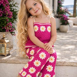 May include: A young girl wearing a bright pink crochet outfit with white and yellow daisy accents. The outfit includes a top and pants. She is wearing gold sandals and has long blonde hair. The background features flowers and a stone structure.