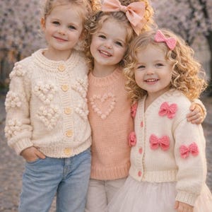 May include: Three young girls in matching outfits, smiling at the camera. One wears a cream cardigan with heart-shaped details, another a pink sweater with a heart, and the third a cream cardigan with pink bows. All have bows in their hair.