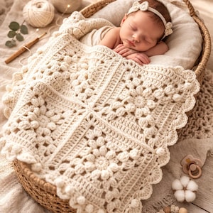 以下が含まれることがあります： クリーム色の手編みの毛布に包まれた、柳のバスケットの中の眠っている赤ちゃん。毛布は花柄で、縁はスカラップ模様です。赤ちゃんはリボン付きのヘアバンドをしています。シーンはニュートラルなトーンのアクセサリーで飾られています。