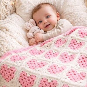 Puede incluir: Un bebé yace en una cuna de macramé color crema, bajo una manta de ganchillo rosa y crema con patrones de corazón. El bebé sostiene un pequeño conejito de ganchillo beige, sonriendo. La manta es un artículo acogedor, hecho a mano.