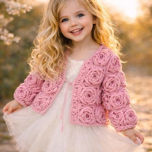 Può includere: Una bambina indossa un vestito bianco e una giacca rosa all'uncinetto decorata con motivi a rosa. La giacca è rosa chiaro e il vestito ha una gonna a strati. La bambina ha i capelli biondi ricci e sorride.