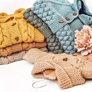 May include: A selection of knitted baby cardigans in shades of mustard yellow, sage green, and teal blue. Each cardigan has a cable knit pattern and heart-shaped wooden buttons. A light tan cardigan is draped over a wooden hanger.