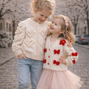 May include: Two young children wearing cream-colored knit cardigans. One cardigan features heart-shaped details, the other has red bow accents. One child wears a pink tulle skirt and the other wears blue jeans. Both children are smiling.