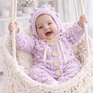 May include: A baby in a lilac knitted romper suit and matching bonnet with bobbles, sitting in a cream macrame swing. The romper has wooden buttons and a ribbon tie. The baby is smiling.