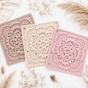 Puede incluir: Tres motivos cuadrados de ganchillo con intrincados patrones florales y puntos de bodoque en relieve. Los motivos son de color malva apagado, crema y rosa suave, cada uno con un diseño central detallado con pétalos radiantes y bordes.