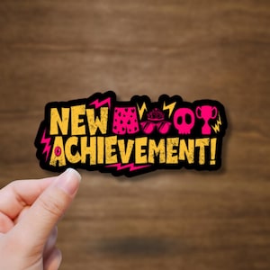 May include: A die-cut sticker featuring the yellow text NEW ACHIEVEMENT! with a black outline and pink lightning bolt accents. The design includes small pink icons of shorts, sunglasses, a skull, and a trophy arranged above the text.