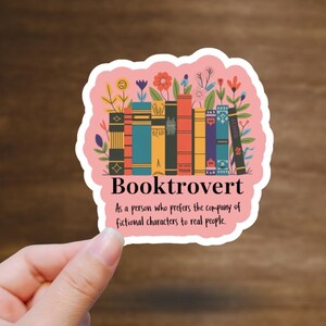 May include: A pink sticker with the word "Booktrovert" and a graphic of colorful books and flowers. The text below reads, "As a person who prefers the company of fictional characters to real people."