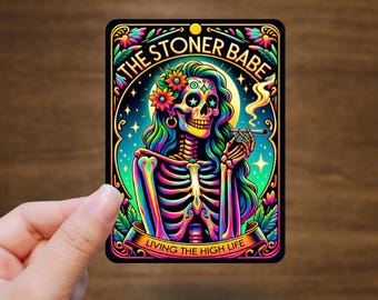 May include: A colorful sticker featuring a skeleton woman with long, wavy hair, a flower crown, and a cigarette. The text reads "THE STONER BABE" and "LIVING THE HIGH LIFE" in a decorative frame with floral accents.