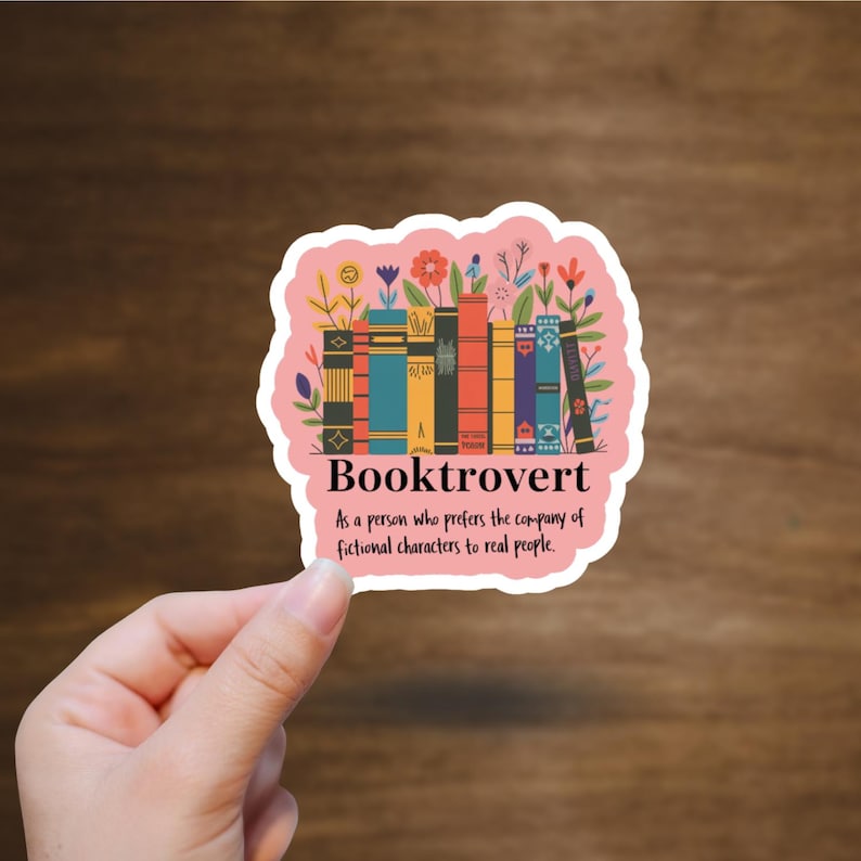 May include: A pink sticker with the word "Booktrovert" and a graphic of colorful books and flowers. The text below reads, "As a person who prefers the company of fictional characters to real people."