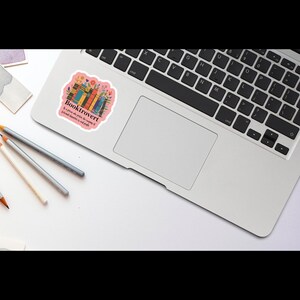 May include: A pink sticker with the word "Booktrovert" and the phrase "A person who prefers the company of fictional characters to real people." The sticker features a colorful illustration of books and flowers, placed on a laptop.