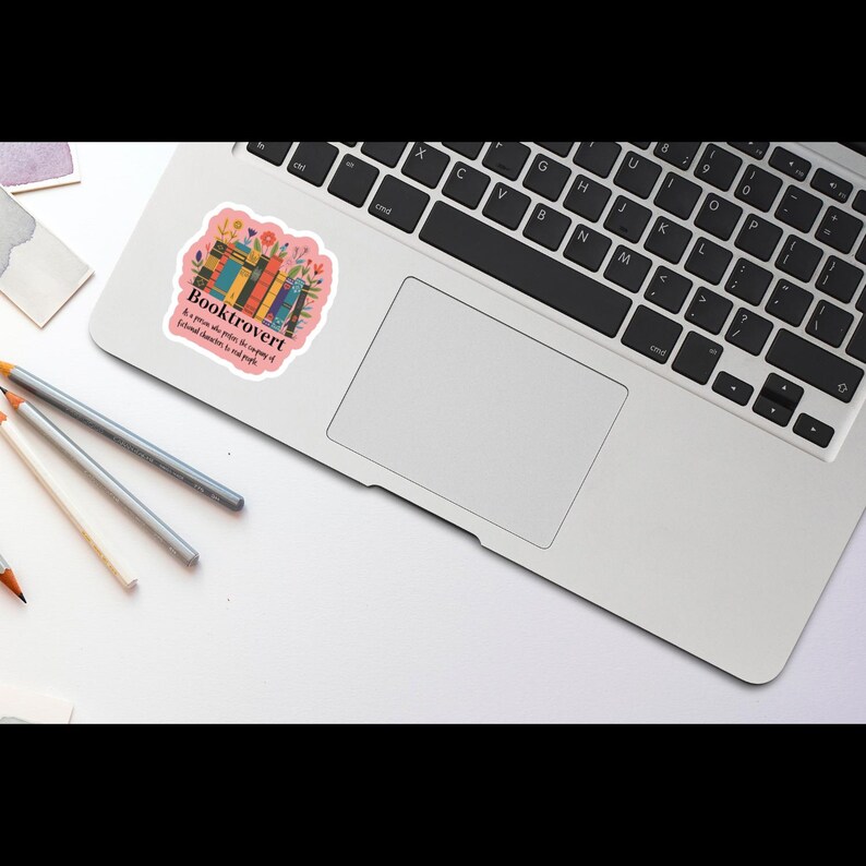 May include: A pink sticker with the word "Booktrovert" and the phrase "A person who prefers the company of fictional characters to real people." The sticker features a colorful illustration of books and flowers, placed on a laptop.