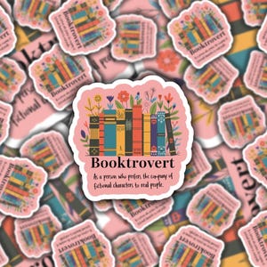 May include: A collection of pink stickers with a central sticker featuring the word "Booktrovert" above a stack of colorful books and flowers. The text "As a person who prefers the company of fictional characters to real people" is also included.