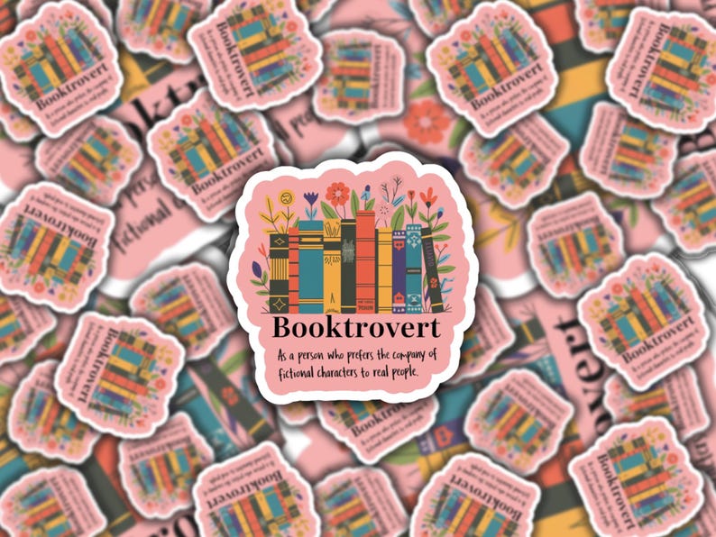 May include: A collection of pink stickers with a central sticker featuring the word "Booktrovert" above a stack of colorful books and flowers. The text "As a person who prefers the company of fictional characters to real people" is also included.