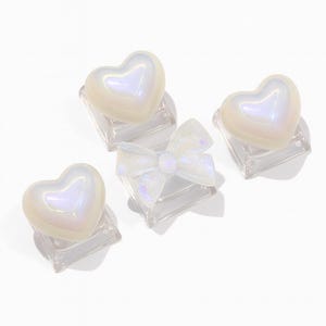 Può includere: Quattro decorazioni iridescenti per nail art. Tre ciondoli a forma di cuore e un ciondolo a forma di fiocco sono montati su basi quadrate trasparenti. I cuori e il fiocco hanno una finitura perlescente, che riflette una gamma di colori. Gli oggetti sono su sfondo bianco.