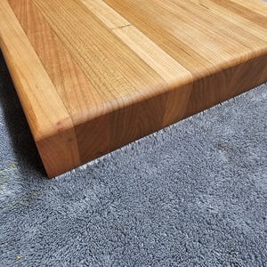 Op de afbeelding: Een vierkant houten snijplank met een lichtbruine afwerking. Het bord is gemaakt van verschillende stukken hout die aan elkaar zijn gezet, met afgeronde randen. De houtnerf is zichtbaar.