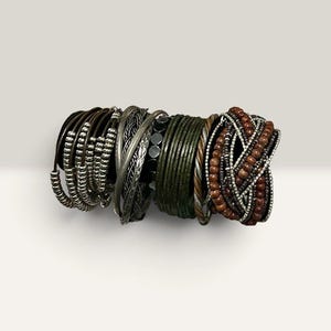 Puede incluir: Un conjunto de pulseras multicapa que presenta una combinación de cordones de cuero marrón oscuro, cuentas metálicas plateadas, alambre plateado trenzado, brazaletes finos de color verde oscuro, alambre de cobre trenzado y cuentas de madera.