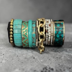 Könnte beinhalten: Ein Stapel Armbänder in verschiedenen Stilen und Farben. Die Armbänder haben türkisfarbene, goldene und Perlenakzente. Einige haben geometrische Muster, andere sind einfarbig. Ein goldenes Kettenarmband ist ebenfalls enthalten.