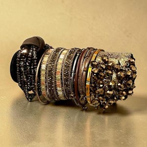 May include: A multi-layered bracelet set featuring a collection of bangles and cuffs in shades of bronze, gold, and dark brown. The set includes bands with faceted dark beads, metallic textured surfaces, and a cluster of dark crystal-like beads.