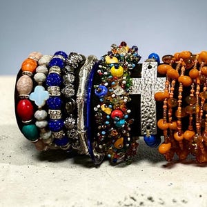 Può includere: Una collezione di bracciali sovrapposti con vari design, tra cui perline in pietra multicolore, un ciondolo a croce azzurro, perline sfaccettate blu scuro con distanziatori argentati, una croce argentata testurizzata e perline arancioni a goccia.