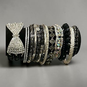 May include: A collection of stacked bracelets featuring a large silver-tone bow encrusted with clear rhinestones, alongside various bands in black and silver tones, including beaded strands, metallic chains, and crystal-embellished designs.
