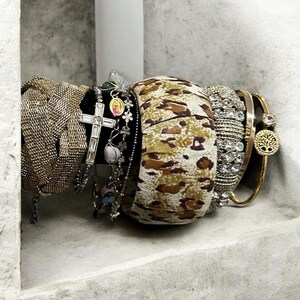 May include: A collection of bracelets including a wide gold-tone mesh cuff, a silver-tone cross charm bracelet, a multi-bead chain, a wide fabric bangle with a tan and brown animal print, a crystal-encrusted band, and a gold-tone tree charm bangle.
