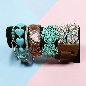 Può includere: Una collezione di bracciali su un supporto nero, tra cui un bracciale con perline turchesi a forma di cuore, un orologio color argento con maglie a cuore, un ampio polsino in filigrana turchese e un bracciale a più fili con piccole perline bianche, blu e rosse.
