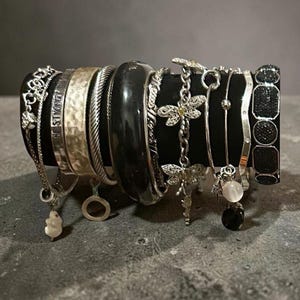 May include: A collection of various bracelets displayed on a black cylindrical stand. The set includes silver-toned chains, a hammered metal cuff engraved with the words BE STRONG, a thick black bangle, a dragonfly charm bracelet, and a black beaded cuff.