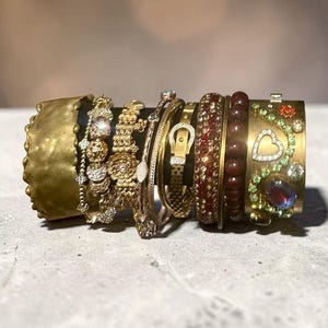 Puede incluir: Una colección de varios brazaletes en tono dorado dispuestos en un soporte. El conjunto incluye un brazalete ancho martillado, varias pulseras de cadena delicadas con detalles florales y de cristal, un brazalete tipo hebilla, una pulsera de cuentas rojas y un brazalete ancho con un motivo de corazón con piedras decorativas verdes y azules.