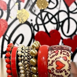May include: A stack of assorted bracelets including a red beaded strand, a solid red bangle, a white pearl strand, a gold chain, a gold beaded strand, another solid red bangle, and a wide cream bangle with red hearts and black text.