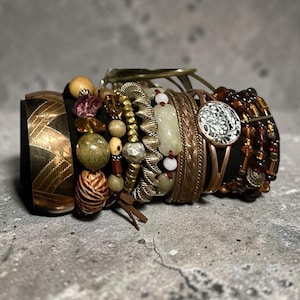 May include: A collection of stacked bracelets featuring a wide etched copper cuff, various beaded strands in shades of brown, green, and cream, a metallic twisted band, a smooth stone segment, and a circular charm with a white patterned inlay.