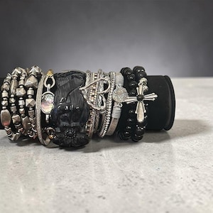 May include: A collection of stacked bracelets featuring various designs including silver-toned metallic beads, a dark grey carved floral cuff, a heart-shaped charm, a circular iridescent inlay, and a black beaded strand with a silver-toned cross charm.