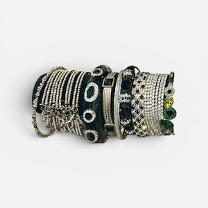 May include: A collection of stacked bracelets featuring various designs, including thin silver-toned chains, a black band with embedded clear stones, a textured silver-toned cuff, and a final bracelet with green and yellow circular accents.