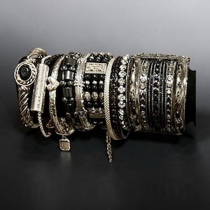 May include: A collection of stacked bracelets featuring silver-toned metal, black beads, and clear crystals. The set includes twisted metal bands, a rectangular charm with engraved text, a heart charm, and several thin, textured bangles.