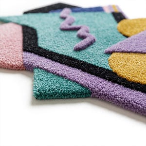 Pode incluir: Close-up de um tapete geom&eacute;trico colorido com uma superf&iacute;cie texturizada e tufada. O tapete apresenta uma combina&ccedil;&atilde;o de cores, incluindo rosa, verde-azulado, roxo, preto e dourado. Um design roxo ondulado est&aacute; no centro. O tapete tem um design moderno e abstrato.