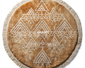 May include: Round, tan rug with a geometric white design and fringe trim. The rug features a repeating pattern of triangles and other shapes. The text "KKASH" is printed in the center of the rug.