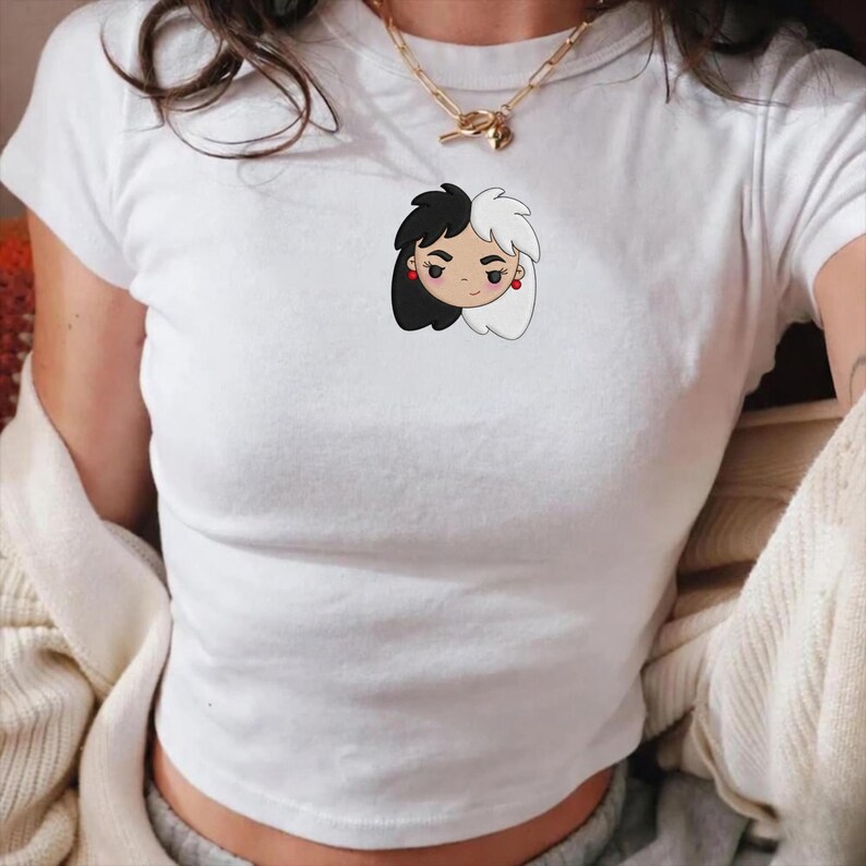 Op de afbeelding: Witte crop top met een cartoon illustratie van een vrouwen gezicht. Het gezicht heeft zwart en wit haar, rode oorbellen en een neutrale uitdrukking. Het shirt is gemaakt van zacht materiaal en heeft korte mouwen.